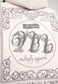 Svetloružová mikina s kapucňou s grafickým potlačou pripomínajúcou poštovú známku. Obsahuje zložitú šedú typografiu, kvetinové akcenty a banner "10 rokov".