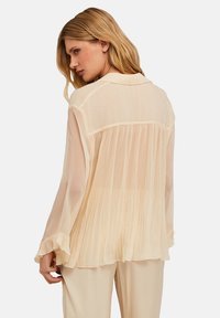Blusa plisada crema con cuello en V, mangas largas y puños con volante. Tejido translúcido con una silueta fluida y espalda texturizada.
