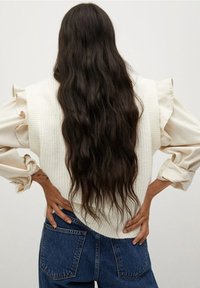 Pull en tricot crème avec manches à volants, assorti à un jean bleu taille haute. Cheveux longs, ondulés et foncés tombant dans le dos.