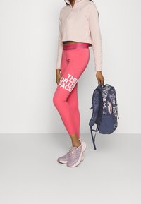 Světle růžový top na čtvrtinový zip, jasně růžové legíny s bílým textem "THE NORTH FACE" a šedé/růžové sportovní boty. Květnatý batoh v ruce.