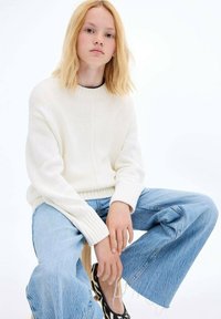 Vit stickad tröja med ribbade manschetter, kombinerad med vida ljusblå jeans. Modellen sitter på en pall och visar avslappnad klädsel.