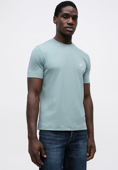 Svijetloplava pamučna majica s okruglim izrezom, kratkim rukavima i malim logotipom na prsima, u kombinaciji s tamnoplavim trapericama.