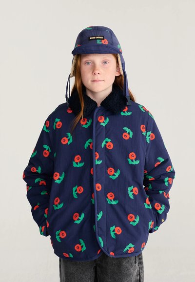 Enfant portant une veste bleu marine et un chapeau assorti avec des motifs floraux rouges vifs et verts, debout les mains dans les poches sur un fond clair.