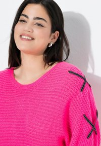 Lächelnde Frau mit dunklem Haar, die silberne Ohrringe und einen leuchtend pinken Strickpullover mit schwarzen gestickten X-Patches auf der Schulter trägt.