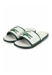 Chanclas blancas con un logo de cocodrilo de goma verde, plantilla texturizada y ribete verde oscuro; suela plana con un acabado acolchado.