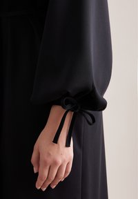 Czarna sukienka z długim, zwiewnym rękawem i marszczonym mankietem związanym tkaną wstążką, prezentująca gładką teksturę i miękki materiał.