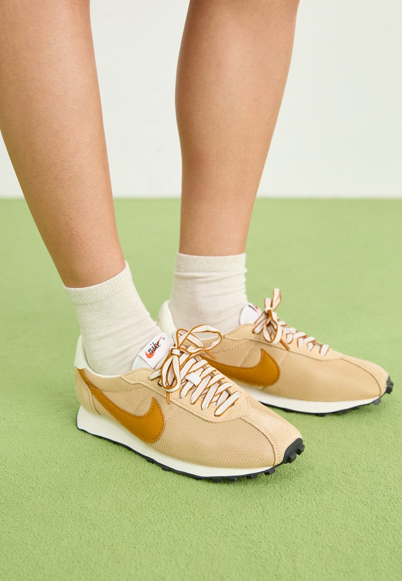 Baskets beiges avec un swoosh orange, tige en mesh et en daim, lacets blancs, semelle noire, associées à des chaussettes beige crème sur une surface verte.
