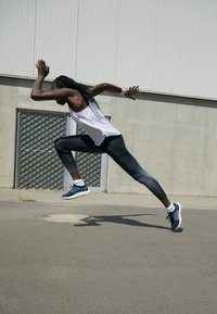 Sportovec v bílém tílku a černých legínách, běžící v vícedobých botách s vlnkovými vzory. V pozadí šedá zeď a okno se vzorem mřížky.