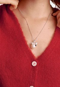 Silberne Halskette mit einem Perlenanhänger und kleinen silbernen Akzenten, getragen über einem roten, strukturierten Cardigan mit Knöpfen.