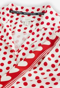 Weißes Hemd mit Kragen, verziert mit roten Punkten, und einem rot-weiß gestreiften Accessoire mit Herzmuster, beschriftet mit "Rockett St George".