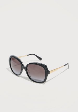 Črne velike sončna očala z gradientnimi temnimi lečami in zlatimi kovinskimi ročaji, ki počivajo na svetli površini pod rahlim kotom.