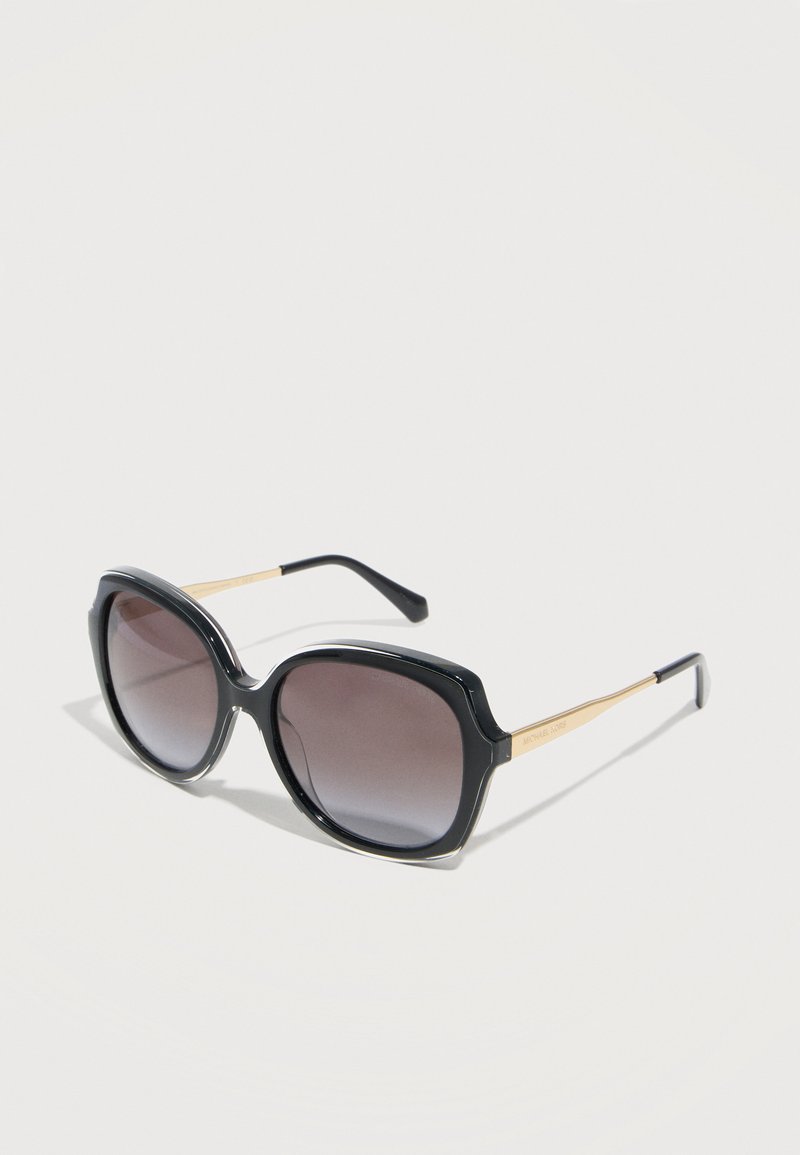 Μεγάλες μαύρες γυαλιά ηλίου με φακούς διαβαθμισμένου σκοταδιού και χρυσά μεταλλικά μπράτσα, ακουμπισμένα σε ανοιχτή επιφάνεια σε ελαφρώς κεκλιμένη γωνία.