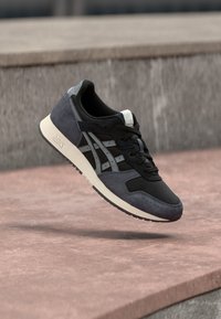 Svart och grå atletisk sko med övre del i mocka och mesh, platta snören och en vit sula. Inkluderar en diagonal randdetalj.