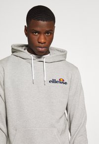 Sudadera gris hecha de tela suave, con un bolsillo frontal, cordones blancos y el logotipo bordado de "ellesse" con un diseño colorido.