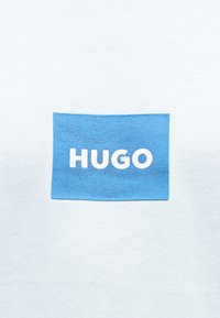 Λευκό ύφασμα με ένα κεντρικά τοποθετημένο μπλε τετράγωνο λάβαρο που έχει τη λέξη «HUGO» σε έντονα λευκά κεφαλαία γράμματα.