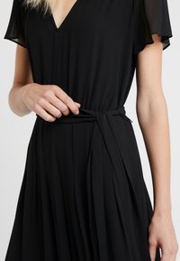 Vestido negro con escote en V y mangas transparentes, que presenta una cintura atada y una falda plisada y fluida. Textura de tela suave.