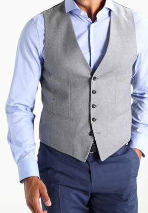 Mann trägt ein hellblaues Hemd, eine graue, geknöpfte Weste und dunkelblaue Hose, mit einer Hand in der Tasche.