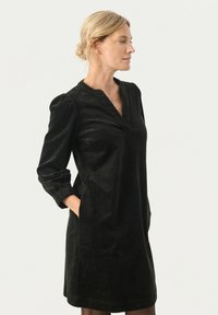 Robe en velours côtelé noir avec un décolleté en V, des manches longues et des poignets élastiques. Chaque côté a des poches ; la texture est douce et côtelée.