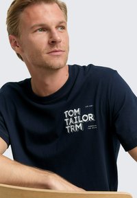 Tamsiai mėlyna medvilninė marškinėliai trumpomis rankovėmis, su baltu grafinio teksto dizainu, kuriame užrašyta "TOM TAILOR" ir šūkis "APIMK LAISVĘ."