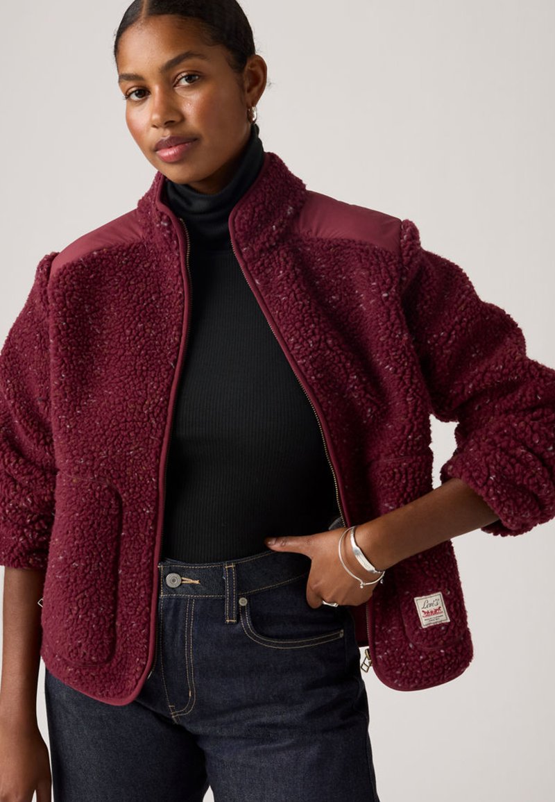 Burgundy fleece jacket with textured, fluffy material, zip closure, and contrasting smooth shoulder panels. Accompanied by dark jeans.