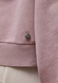 Gros plan de l’ourlet d’un sweat-shirt rose poussiéreux avec un poignet côtelé et un petit emblème rond argenté de la marque attaché près de la couture.