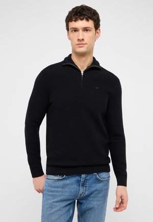 Junger Mann trägt einen schwarzen Halb-Reißverschluss-Pullover und blaue Jeans und steht vor einem schlichten hellen Hintergrund.