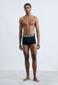 Μαύρες μποξερές Calvin Klein με άσπρη ζώνη λογότυπου. Το μοντέλο στέκεται όρθιο, δείχνοντας την εφαρμογή και τον σχεδιασμό σε ανοιχτό φόντο.