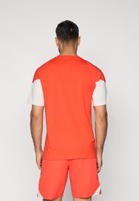 Piros sportpóló rövid ujjakkal, fehér díszítéssel a vállakon és a háton. Sima textúra, egyszerű dizájn, és klasszikus szabás.