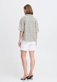 Camicia a maniche corte con bottoni, caratterizzata da righe verticali in beige, verde e rosso, con un orlo leggermente curvo, abbinata a pantaloni corti bianchi.