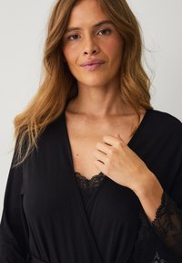 Robe noire de style kimono en tissu doux, avec des détails en dentelle sur les poignets et une bordure en dentelle assortie en dessous. À nouer à la taille.