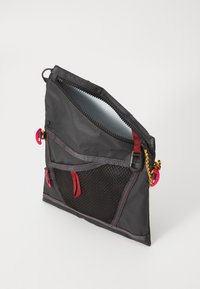 Borsa nera in nylon con tasca a rete, dettagli rossi e chiusura con cerniera; presenta accessori colorati e una superficie testurizzata sulla maniglia.