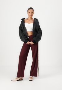 Veste bomber noire, top court blanc, pantalons larges bordeaux avec une rayure beige sur le côté, et des chaussures slip-on beiges avec des strapping. Textures lisses.