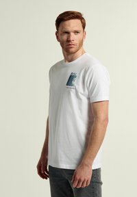 Camiseta blanca de algodón con cuello redondo y mangas cortas, que presenta un diseño impreso en el lado izquierdo del pecho en azul y verde. Ajuste clásico.