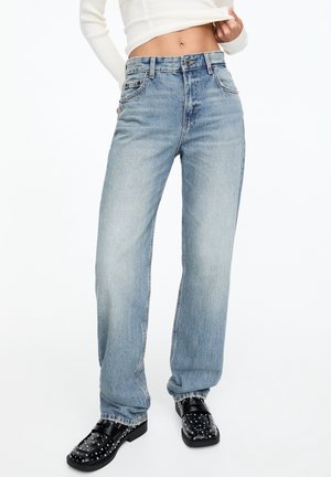 Frau trägt hellblaue High-Waist-Jeans, weißen kurzen Pullover und schwarze Loafers mit Nieten vor einem einfarbigen weißen Hintergrund.