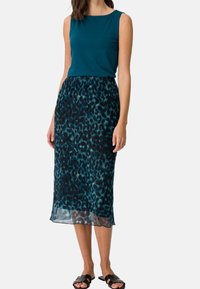 Teal bezrękawnik w połączeniu z półdługą, przezroczystą spódnicą z czarnym i turkusowym wzorem w panterkę; dopełnione czarnymi sandałami.