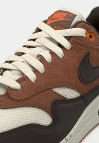 Close-up de um sneaker Nike Air Max marrom, preto e branco com atacadores creme e logótipo laranja na lingueta.