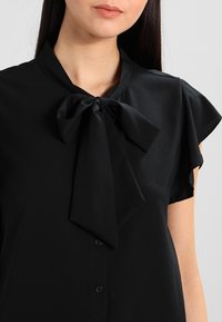 Blusa negra con mangas cortas con volantes y un lazo grande en el cuello. Hecha de tela suave con botones delanteros y una textura lisa.