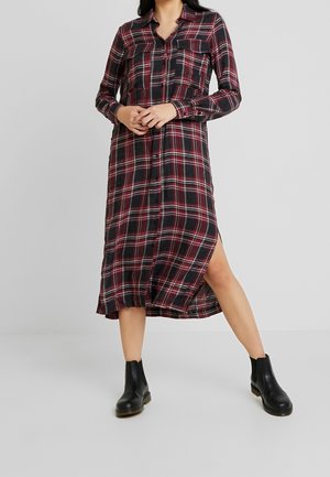 Femme portant une robe midi à manches longues rouge et noire à carreaux, boutonnée avec des fentes latérales et des bottines noires, debout jambes écartées.