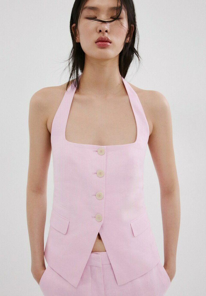 Donna con capelli lisci e scuri che indossa un gilet rosa chiaro senza maniche con bottoni e pantaloni abbinati su uno sfondo neutro.