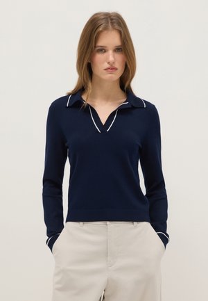 Frau mit glatten hellbraunen Haaren, die einen marineblauen Langarm-Polopullover mit weißem Besatz und beigefarbenen Hosen trägt, mit den Händen in den Taschen vor neutralem Hintergrund.