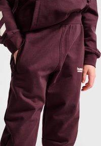 Close-up van een persoon die bordeauxrode trainingsbroek en hoodie draagt met hand in de zak, waarbij het hummel sportkledinglogo op de broek zichtbaar is.