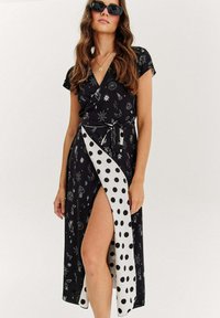 Černé zavinovací šaty s hvězdným vzorem, krátkými rukávy a bočním panelem s puntíkovým vzorem. Lehce průsvitný materiál, s páskem na zavázání v pase a bočním rozparem.