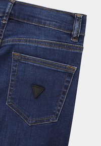 Dark blue denim jeans featuring a rear pocket with a black triangular logo patch and contrasting yellow stitching.