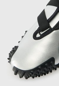 Sapatilha atlética metálica prateada com uma superfície texturizada, picos de borracha pretos na sola e uma faixa preta para um ajuste seguro.