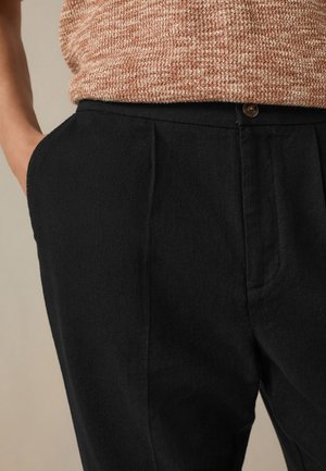 Personne portant un pantalon noir plissé avec des poches latérales et un haut en maille beige texturé rentré, main reposant dans la poche.