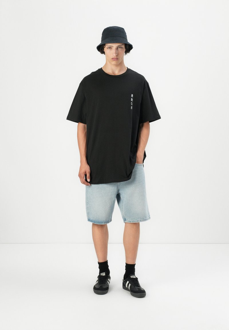 Schwarzes, oversized T-Shirt mit seitlichem Logo-Print, kombiniert mit hellen Denim-Shorts. Das Model trägt einen schwarzen Bucket Hat und schwarze Sneakers.