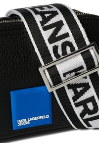 Schwarze Lederhandtasche mit einem blauen rechteckigen Logo, einem breiten verstellbaren Träger mit schwarz-weißer Aufschrift und einer silbernen Schnalle.