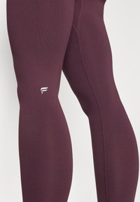 Bordeaux leggings gemaakt van rekbaar materiaal, met een gladde textuur, gevormde naden en een klein wit logo op het onderbeen.