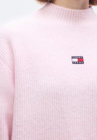 Pulover roz din tricot cu dungi, cu guler înalt și o mică etichetă dreptunghiulară cu logo-ul „TOMMY JEANS” în albastru închis și roșu, poziționată pe piept.