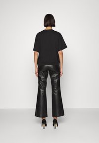 Mujer de espaldas a la cámara, vistiendo una blusa negra de manga corta, pantalones de cuero negros acampanados y zapatos de tacón negro.